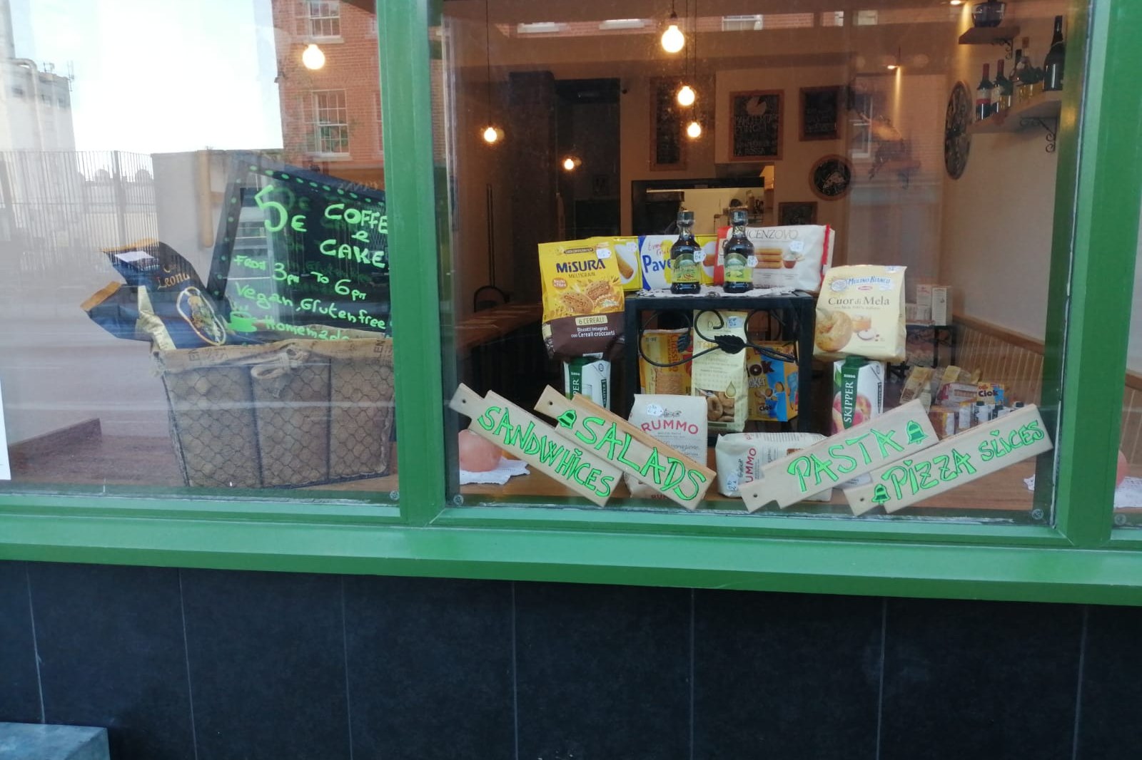The shopfront window at Bell Pesto Café, with green-painted signage and hand-painted boards for sandwiches, salads, pasta and pizza slices.